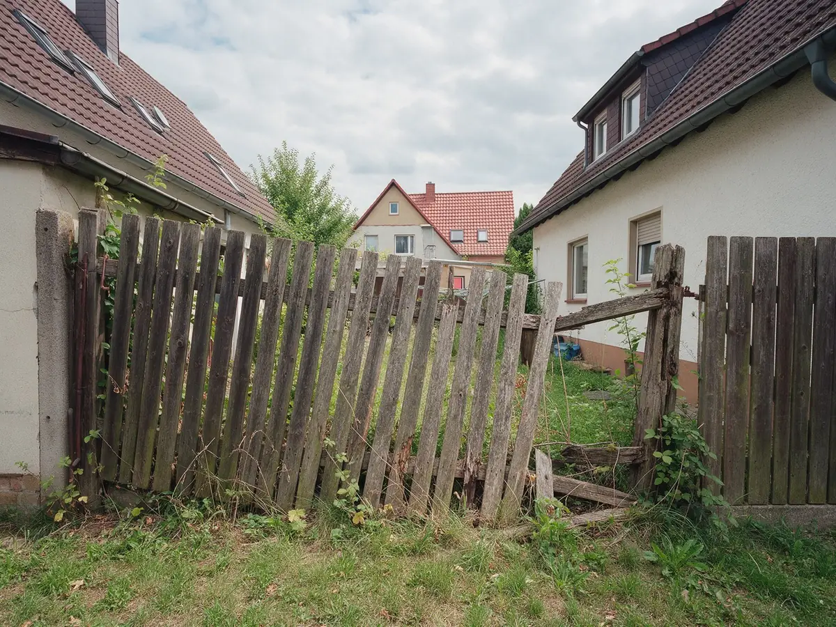 KI-generiertes Beispiel, Vorher: Schiefer, morscher Holzzaun mit fehlenden Latten an einer Grundstücksgrenze